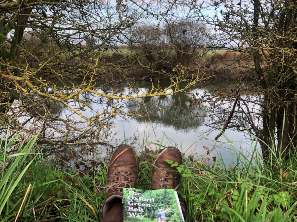 a view of a river with foliage around, with a view of the photographer's feet, resting on the riverbank, and the Oxford Green Belt Way guidebook on her lap