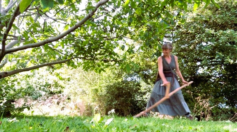 A woman scything wearing a long skirt and smiling
