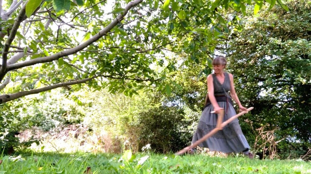 A woman scything wearing a long skirt and smiling