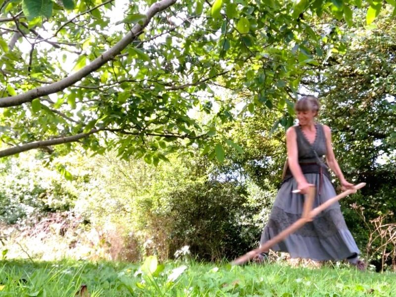 A woman scything wearing a long skirt and smiling