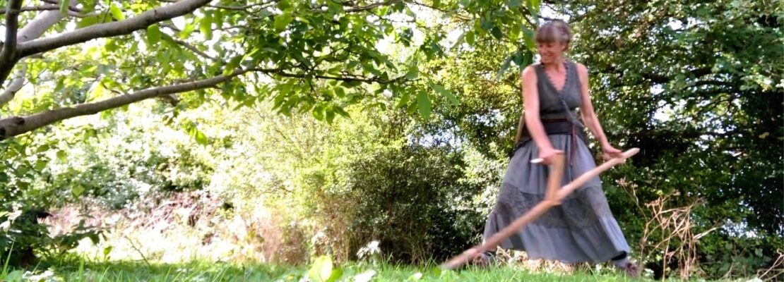 A woman scything wearing a long skirt and smiling