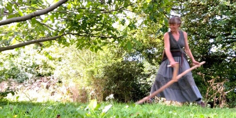 A woman scything wearing a long skirt and smiling
